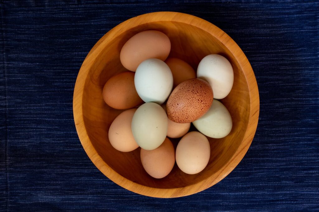 pexels photo 9331860 A variety of colorful chicken eggs arranged in a wooden bowl on a dark blue background.